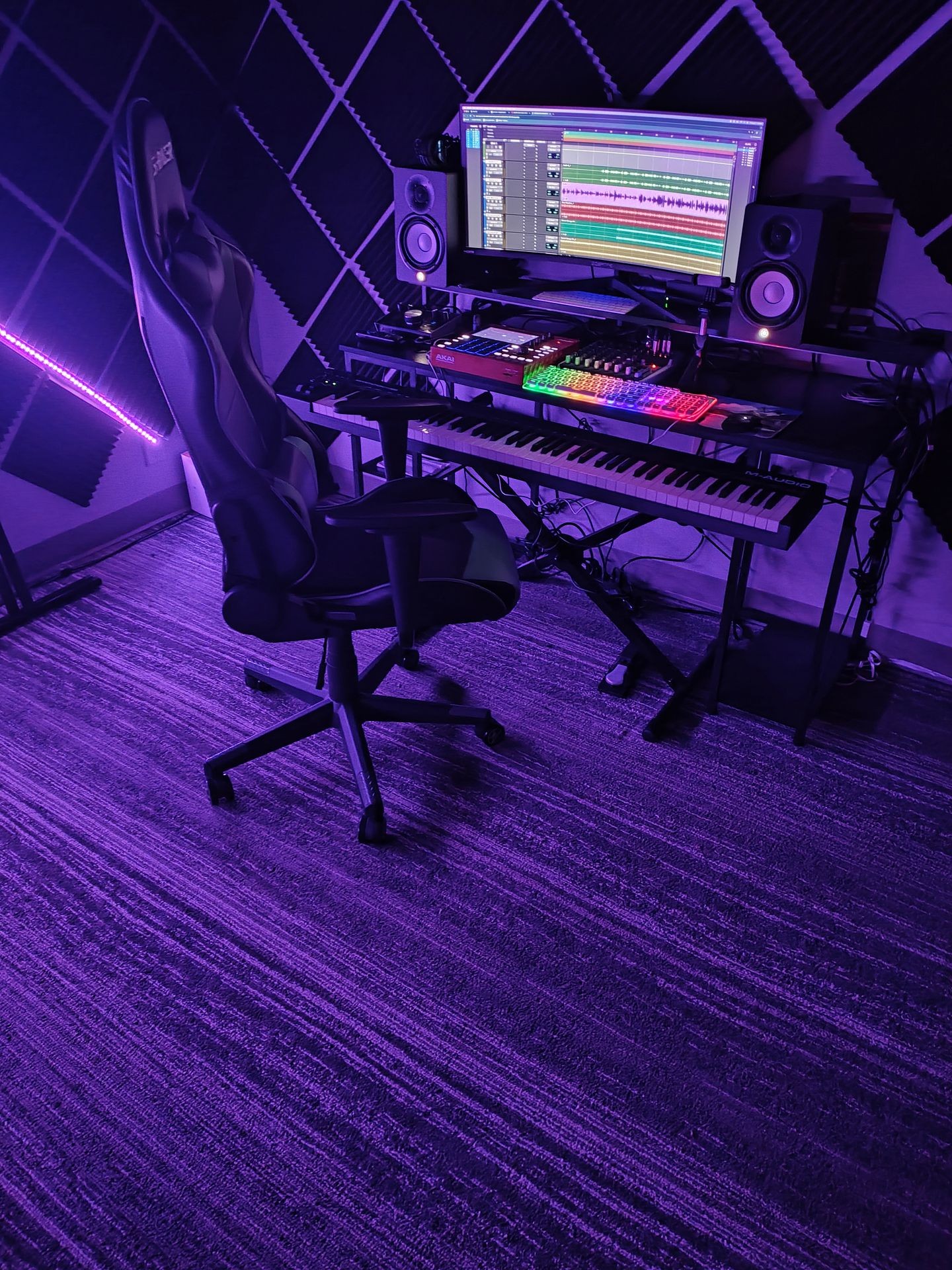 Music production studio with monitors, keyboard, and gaming chair under purple lighting.